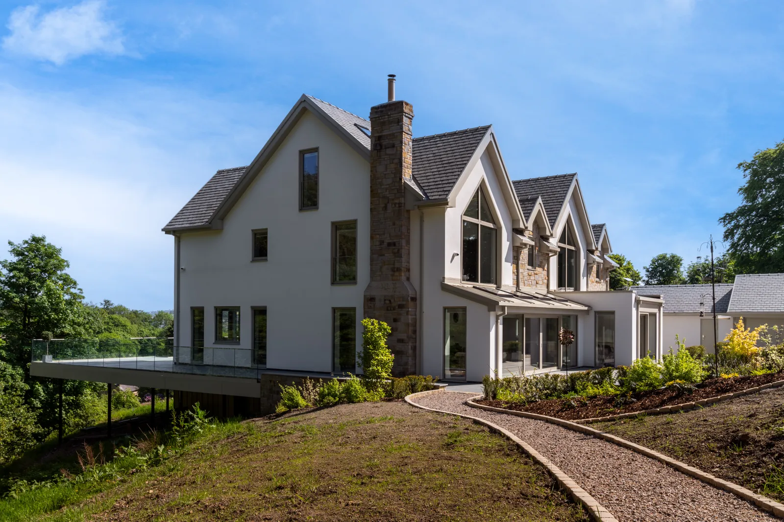 Side exterior of a modern white render and stone house with pitched gables and landscaped garden