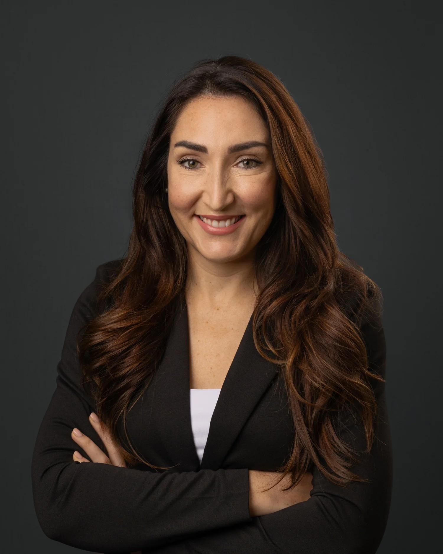 Corporate headshot of a woman with dark wavy hair in a black blazer on a grey background