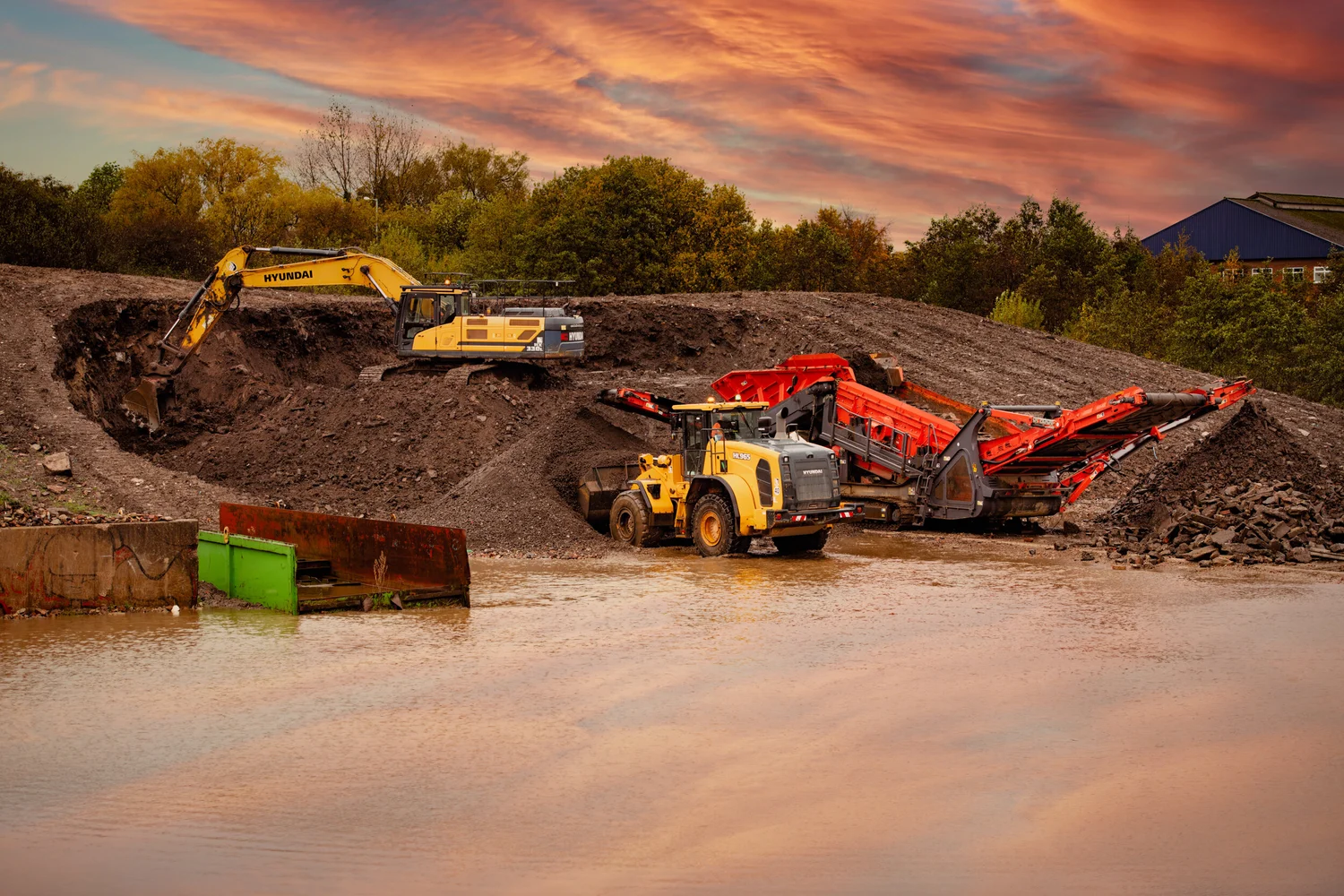 Excavator and wheel loader working beside a mobile crushing rig at sunset on a recycling site